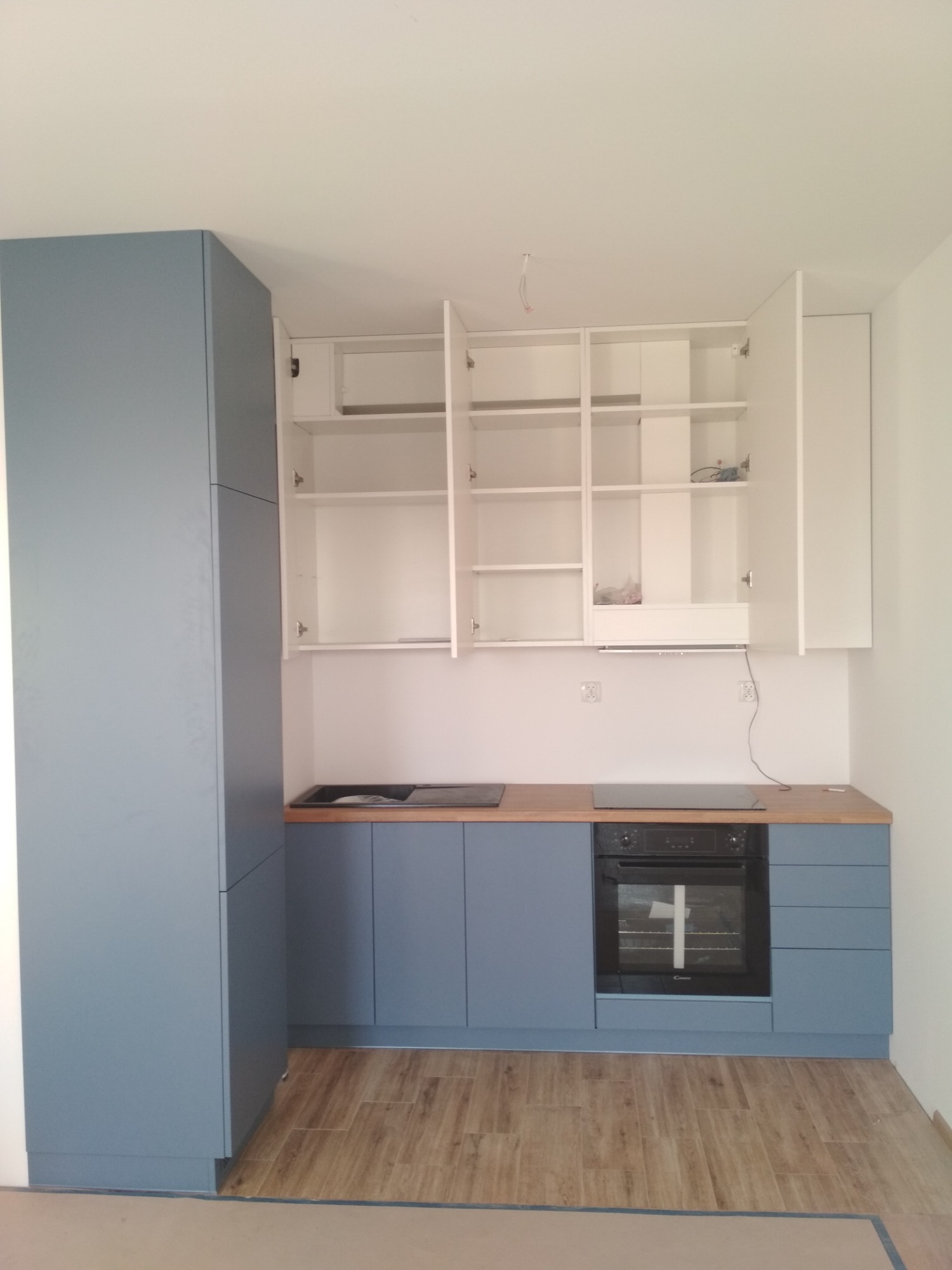 Niebiesko-biała zabudowa kuchenna z otwartymi szafkami wiszącymi, drewnianym blatem i płytą indukcyjną, widok na wnętrze piekarnika.