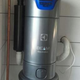 DECOR Damian Marek - Szara jednostka centralna odkurzacza BEAM Electrolux zamontowana na ścianie z białych płytek, widoczny panel sterowania z niebieskim podświetleniem i podłączenia rur.