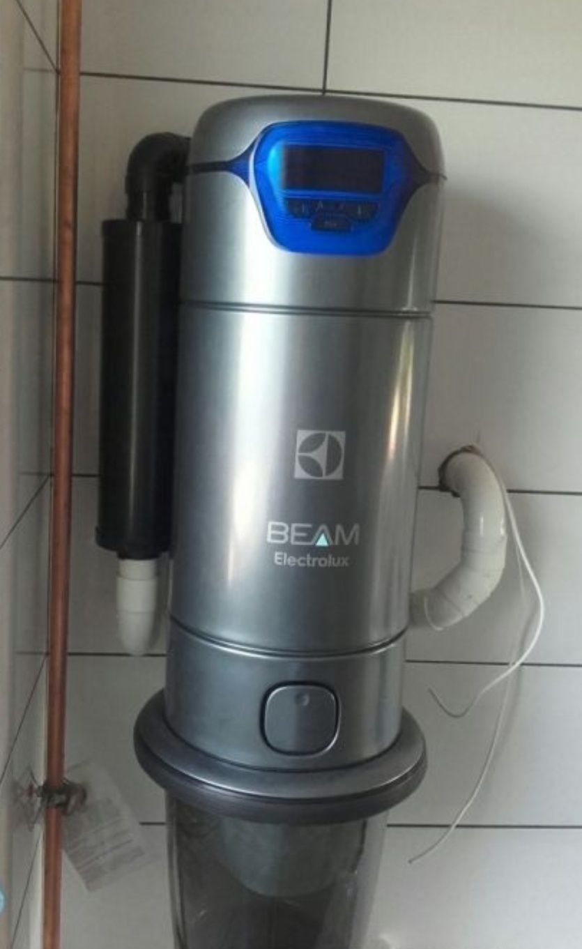 Szara jednostka centralna odkurzacza BEAM Electrolux zamontowana na ścianie z białych płytek, widoczny panel sterowania z niebieskim podświetleniem i podłączenia rur.