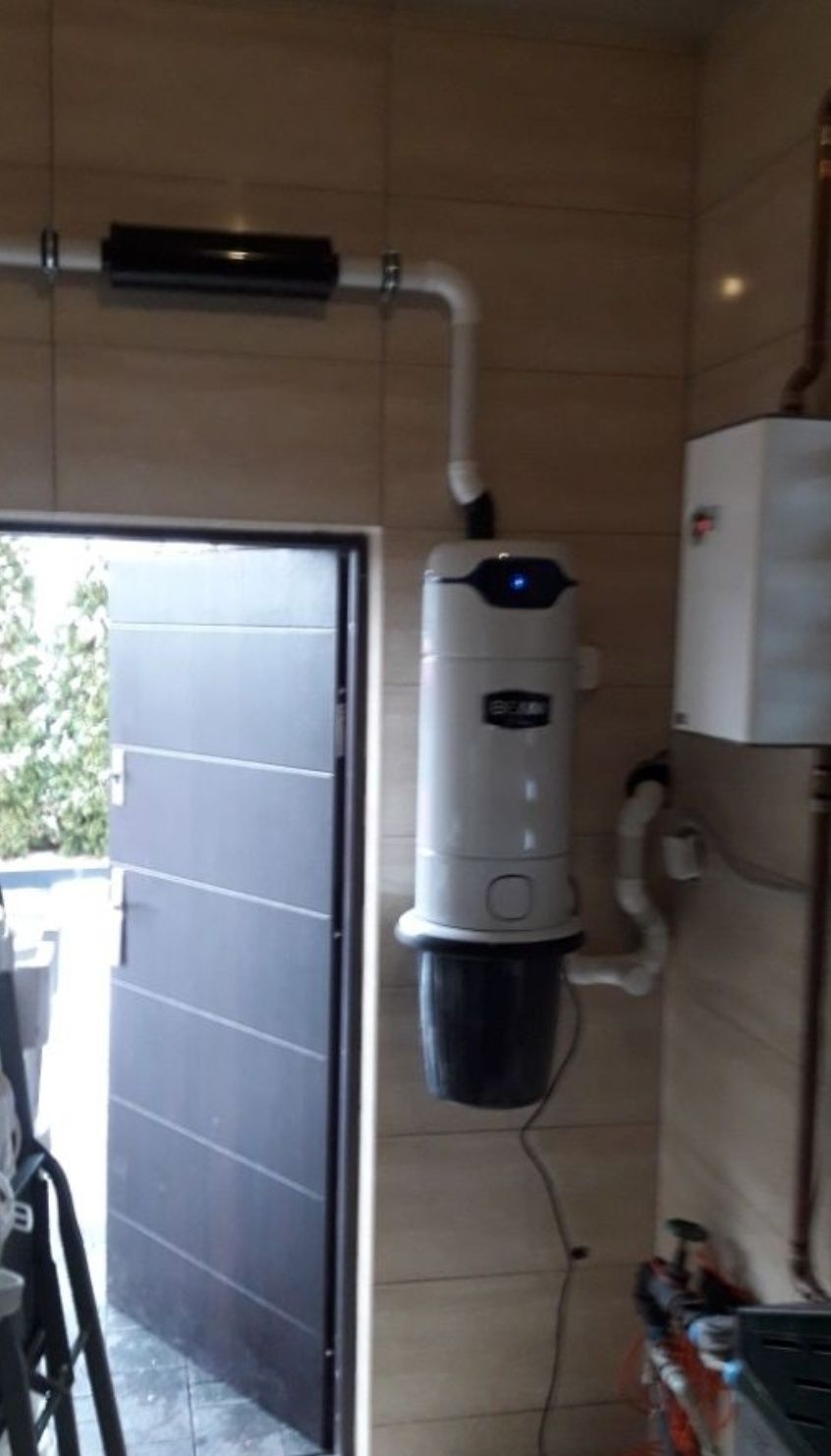 Centralny odkurzacz BEAM z podłączonymi rurami wentylacyjnymi, zamontowany na kafelkowanej ścianie w pobliżu otwartych drzwi garażowych.