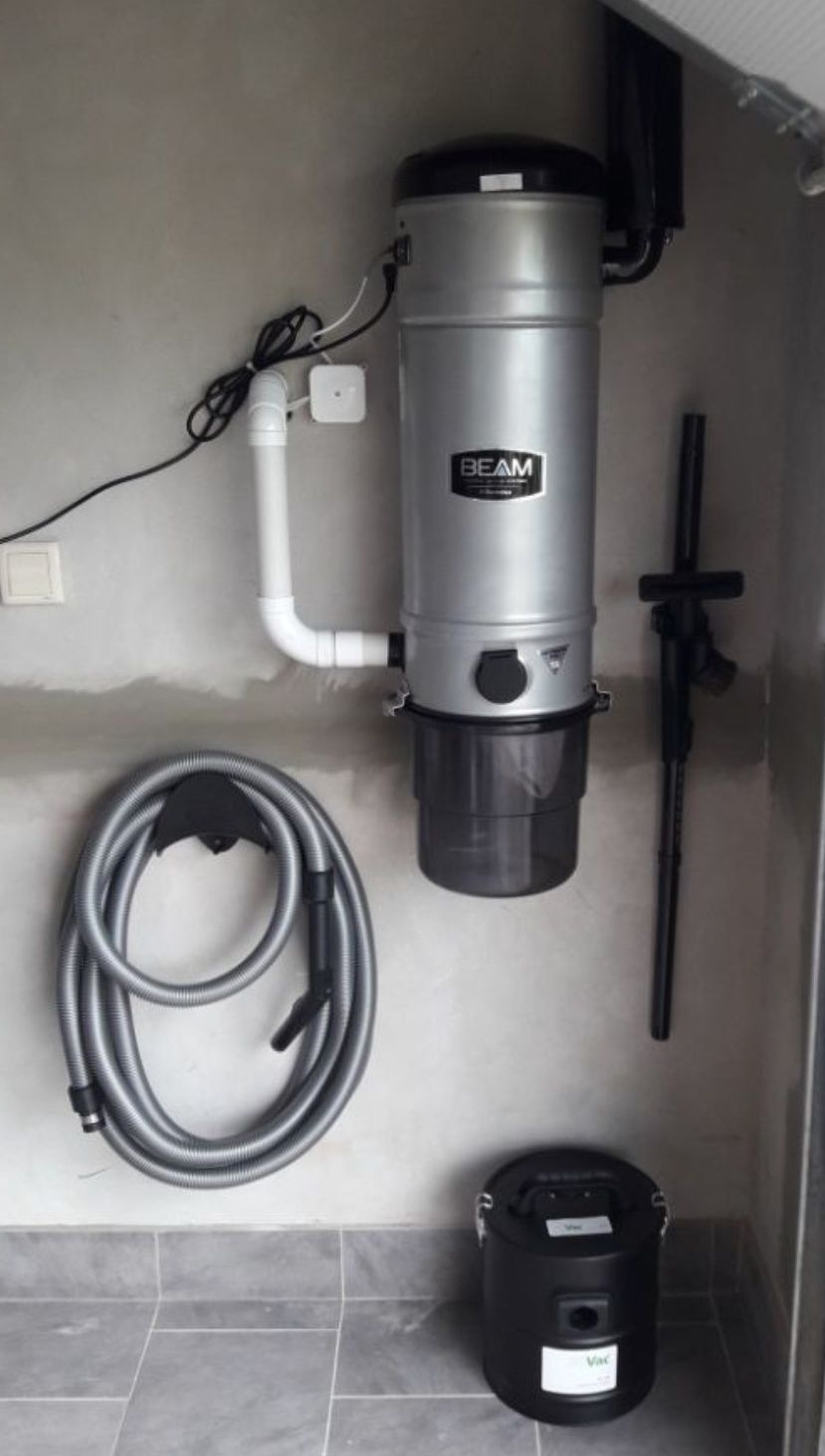 Centralny odkurzacz BEAM zamontowany na szarej ścianie, z widocznym wężem ssącym na uchwycie i pojemnikiem na kurz na podłodze z szarych płytek.