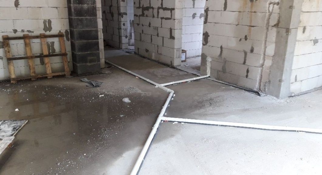 Instalacja hydrauliczna w surowym stanie: białe rury PVC ułożone na mokrej, betonowej posadzce w budynku z białych bloczków, widoczna drewniana paleta.
