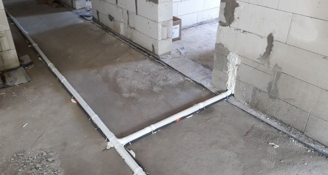 Białe rury instalacyjne ułożone w bruzdach w podłodze surowego budynku z betonu komórkowego, widoczne łączenia i fragmenty czarnych rur, przygotowane do zalania wylewką.