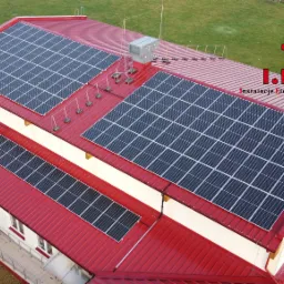 Widok z góry na budynek z czerwonym dachem, na którym zamontowano panele fotowoltaiczne; na dachu widoczny także odgromnik oraz logo firmy zajmującej się instalacjami elektrycznymi.