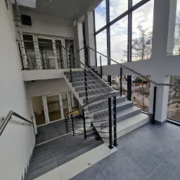 Wnętrze budynku z dwubiegowymi schodami wykończonymi szarą płytką i metalową balustradą z poziomymi prętami, widok z góry na dół, duże okno z widokiem na zewnątrz.