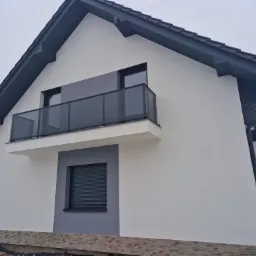 Nowoczesny dom z balkonem zabezpieczonym szklaną balustradą w kolorze grafitowym, widok z boku, z fragmentem ośnieżonego terenu.