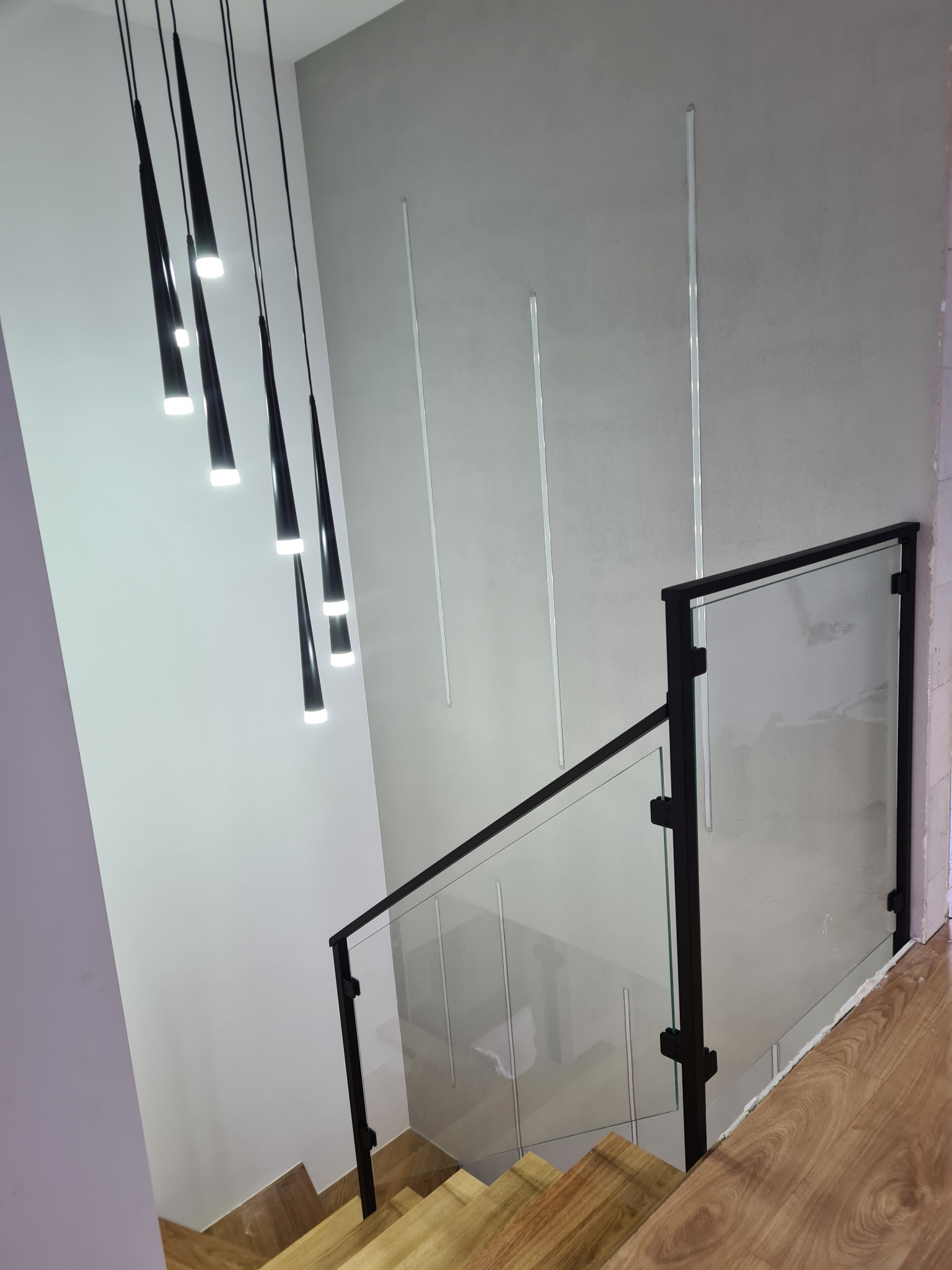 Wewnętrzna balustrada schodowa z czarnymi profilami i przezroczystym szkłem hartowanym, oświetlona wiszącymi lampami w kształcie stożków, widok z góry na drewniane schody.