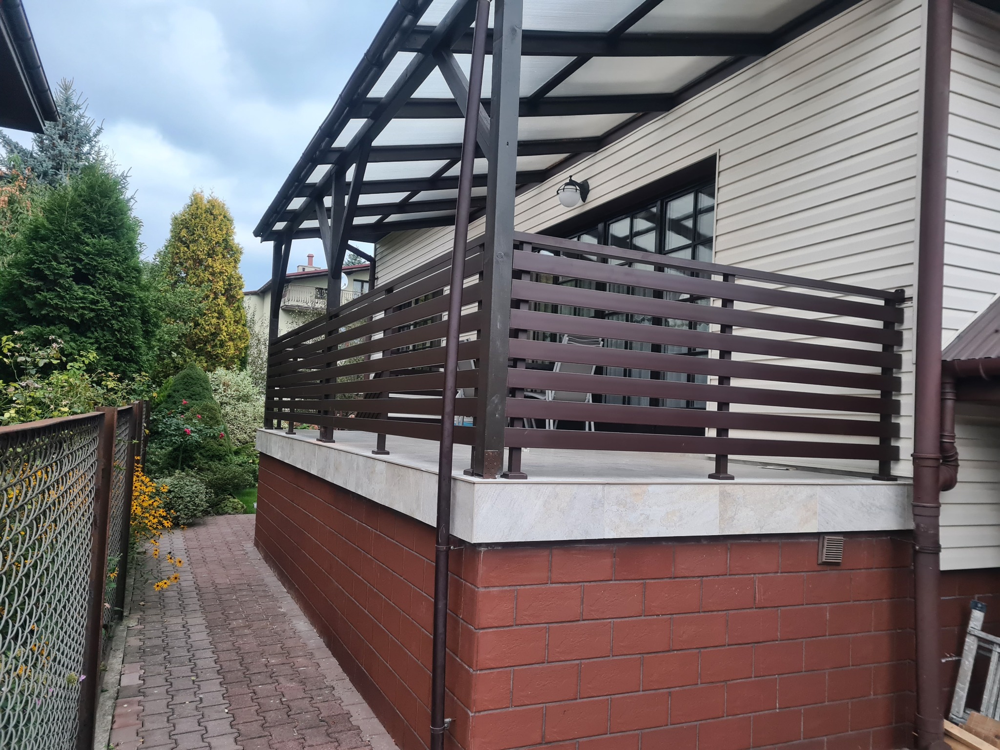 Zadaszony taras z brązową, poziomą balustradą, wykończony cegłą i jasnymi płytkami, widok z boku ścieżki z kostki brukowej.