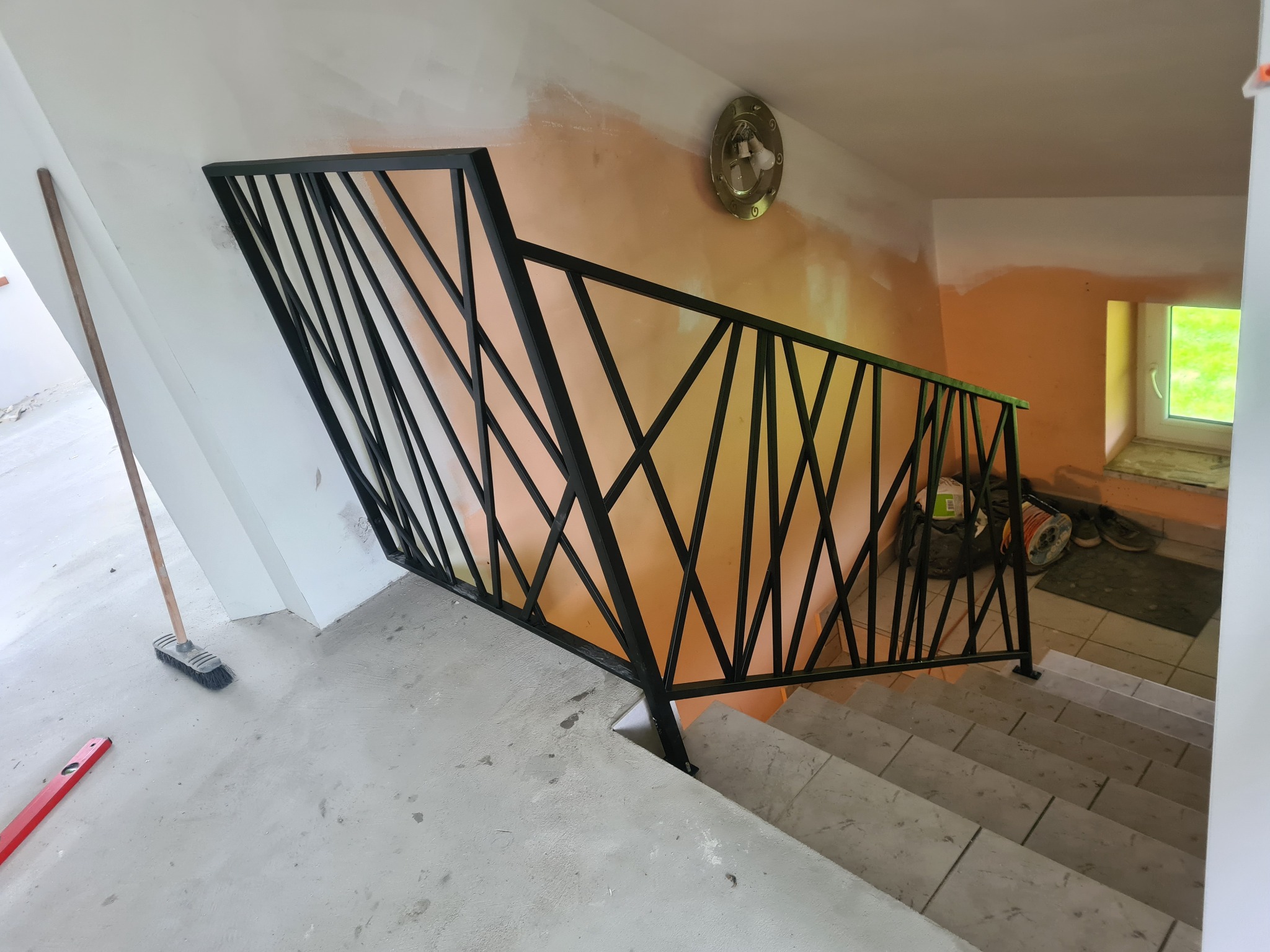 Nowoczesna, czarna balustrada schodowa z geometrycznym wzorem, widoczna na tle pomarańczowej ściany w trakcie malowania i schodów wyłożonych płytkami. Na podłodze leżą narzędzia: poziomica i zmiotka.