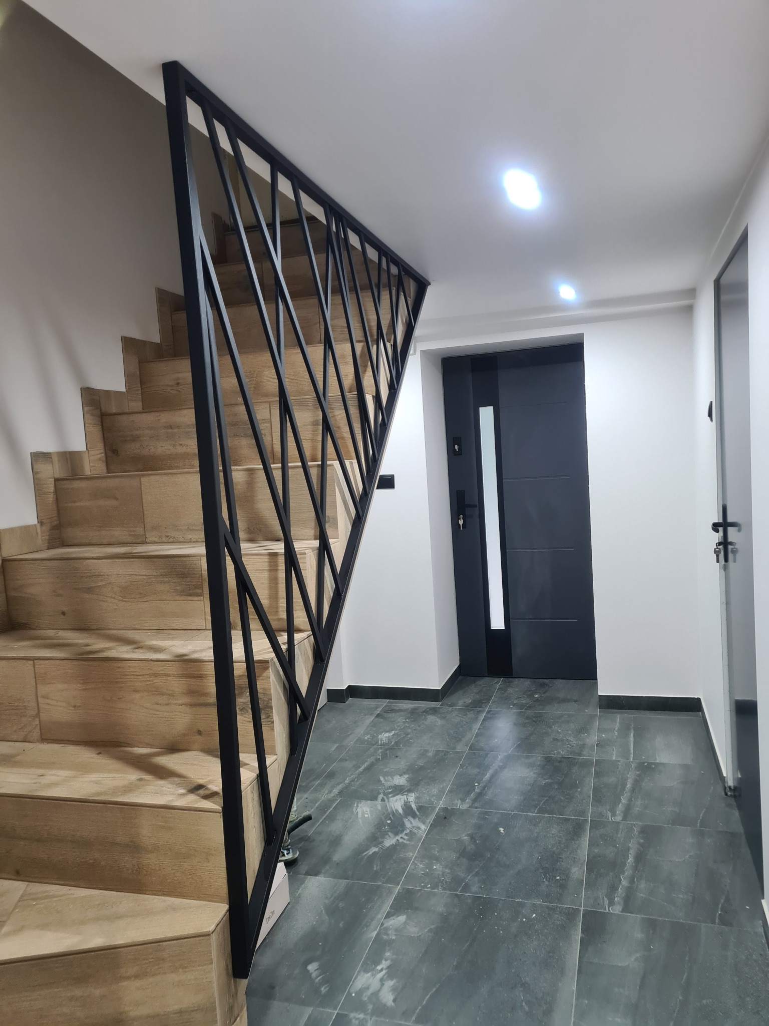 Schody wyłożone płytkami imitującymi drewno, zabezpieczone czarną, metalową balustradą o geometrycznym wzorze, w tle ciemne drzwi i szara podłoga.