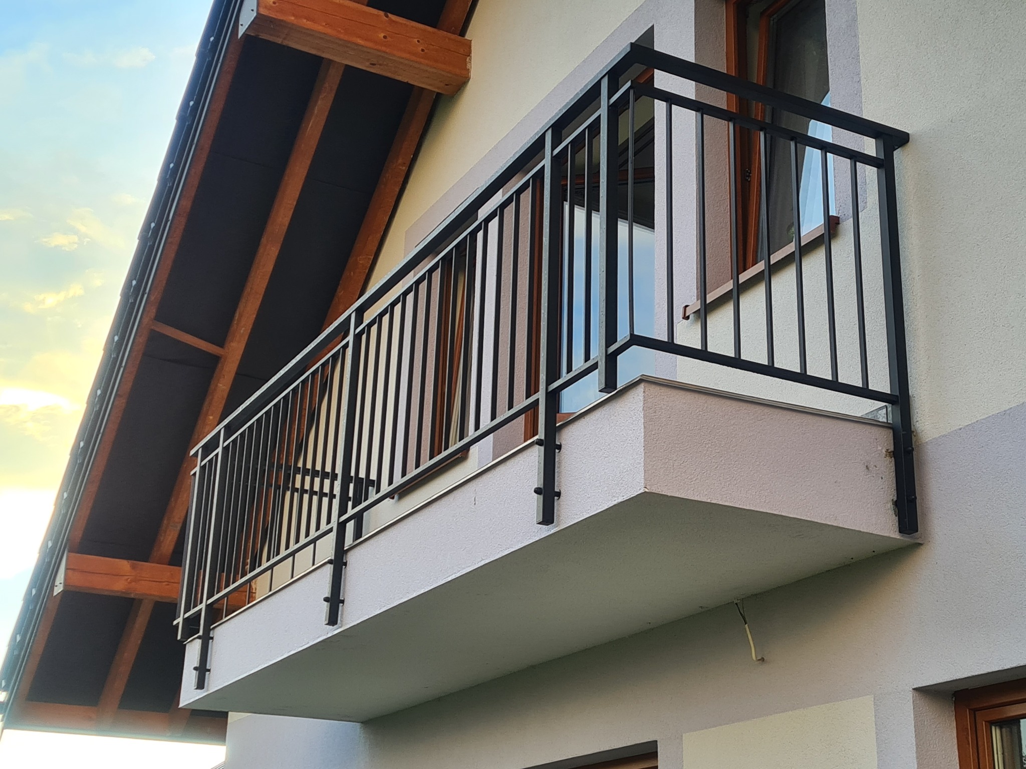 Nowoczesna, czarna balustrada balkonowa z pionowymi prętami, zamontowana na balkonie z szarą elewacją budynku i drewnianą konstrukcją dachu w tle.