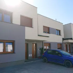 Nowoczesne domy szeregowe z szarymi i białymi elewacjami, brązowymi oknami z roletami, zaparkowany niebieski samochód osobowy, kostka brukowa na podjeździe.