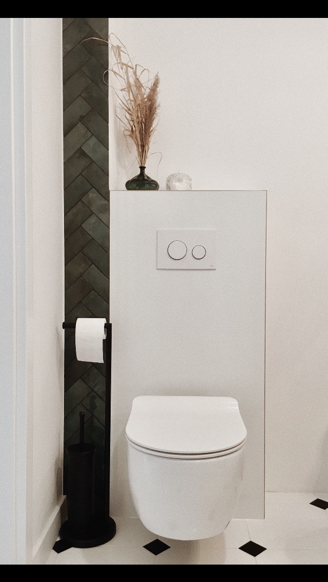 Nowoczesna łazienka z podwieszaną toaletą, zieloną ścianą z kafelków ułożonych w jodełkę, czarnym stojakiem na papier toaletowy i szczotkę oraz dekoracją z suszonych traw w ciemnozielonym wazonie.