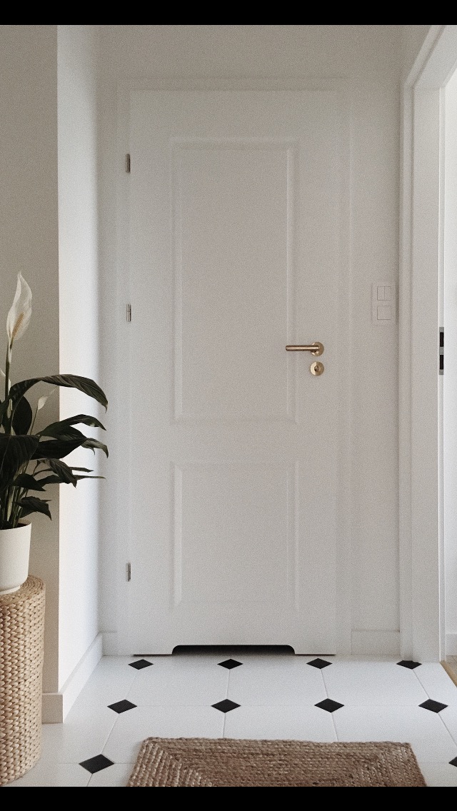 Białe drzwi wewnętrzne z złotą klamką, biała podłoga z czarnymi rombami, wiklinowy stolik z rośliną z białymi kwiatami, minimalistyczny styl.