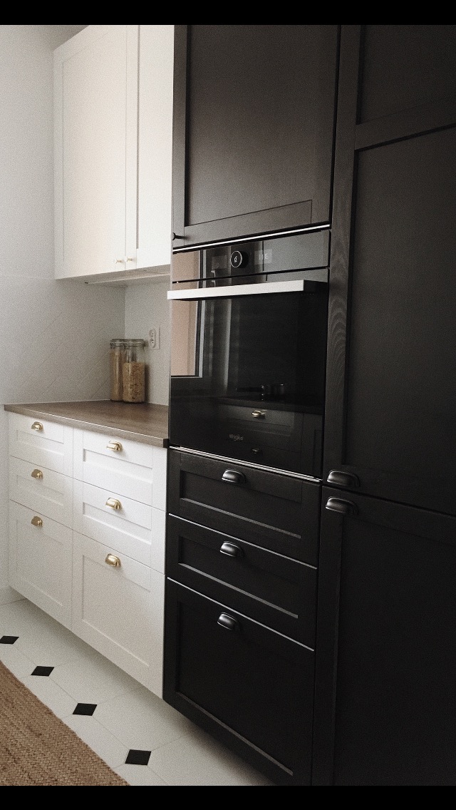 Kontrastowe szafki kuchenne: białe z mosiężnymi uchwytami po lewej, czarne z ciemnymi uchwytami po prawej, z wbudowanym piekarnikiem Whirlpool i blatem z efektem drewna.