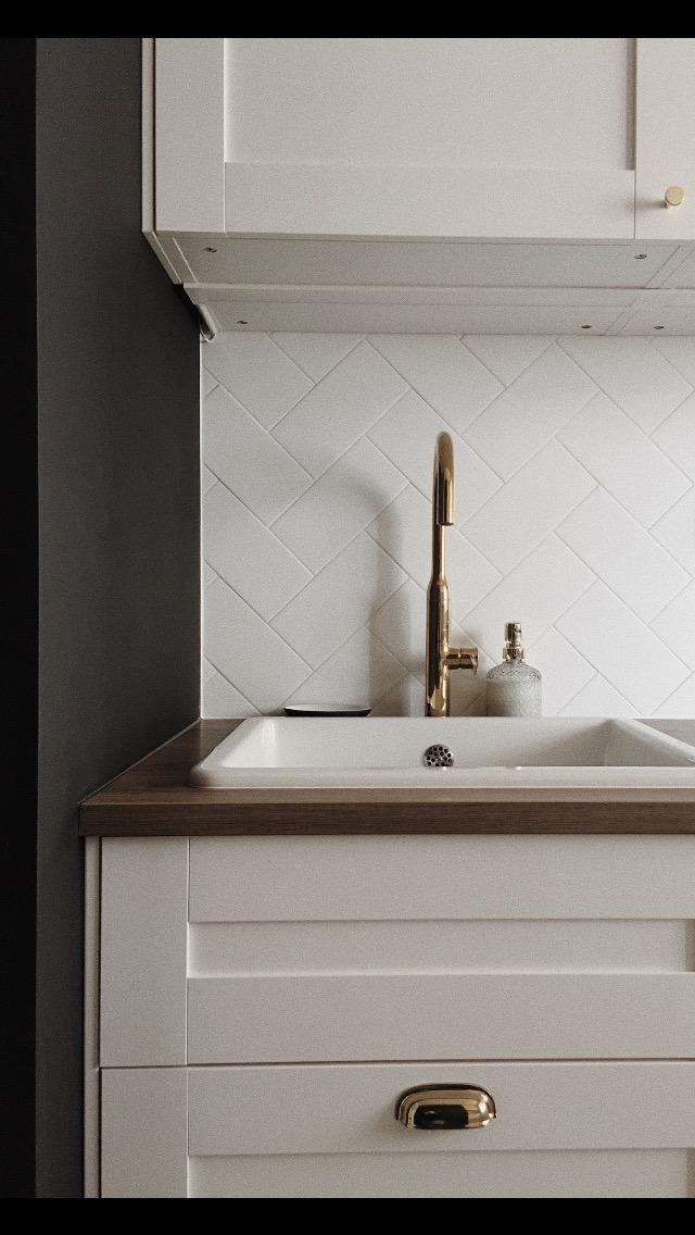 Nowoczesna łazienka: biała, podwójna umywalka z baterią w kolorze złotym, białe płytki ułożone w jodełkę na ścianie, drewniany blat i białe szafki z frezowanymi frontami.