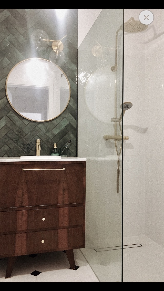 Nowoczesna łazienka z drewnianą szafką pod umywalką, okrągłym lustrem w złotej ramie, zielonymi płytkami ułożonymi w jodełkę i kabiną prysznicową typu walk-in ze złotą armaturą.