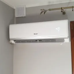 Biały klimatyzator marki GREE zamontowany na szarej ścianie, obok okna z drewnianą ramą i zasłoniętymi roletami, z wyświetlaną temperaturą 20 stopni Celsjusza.