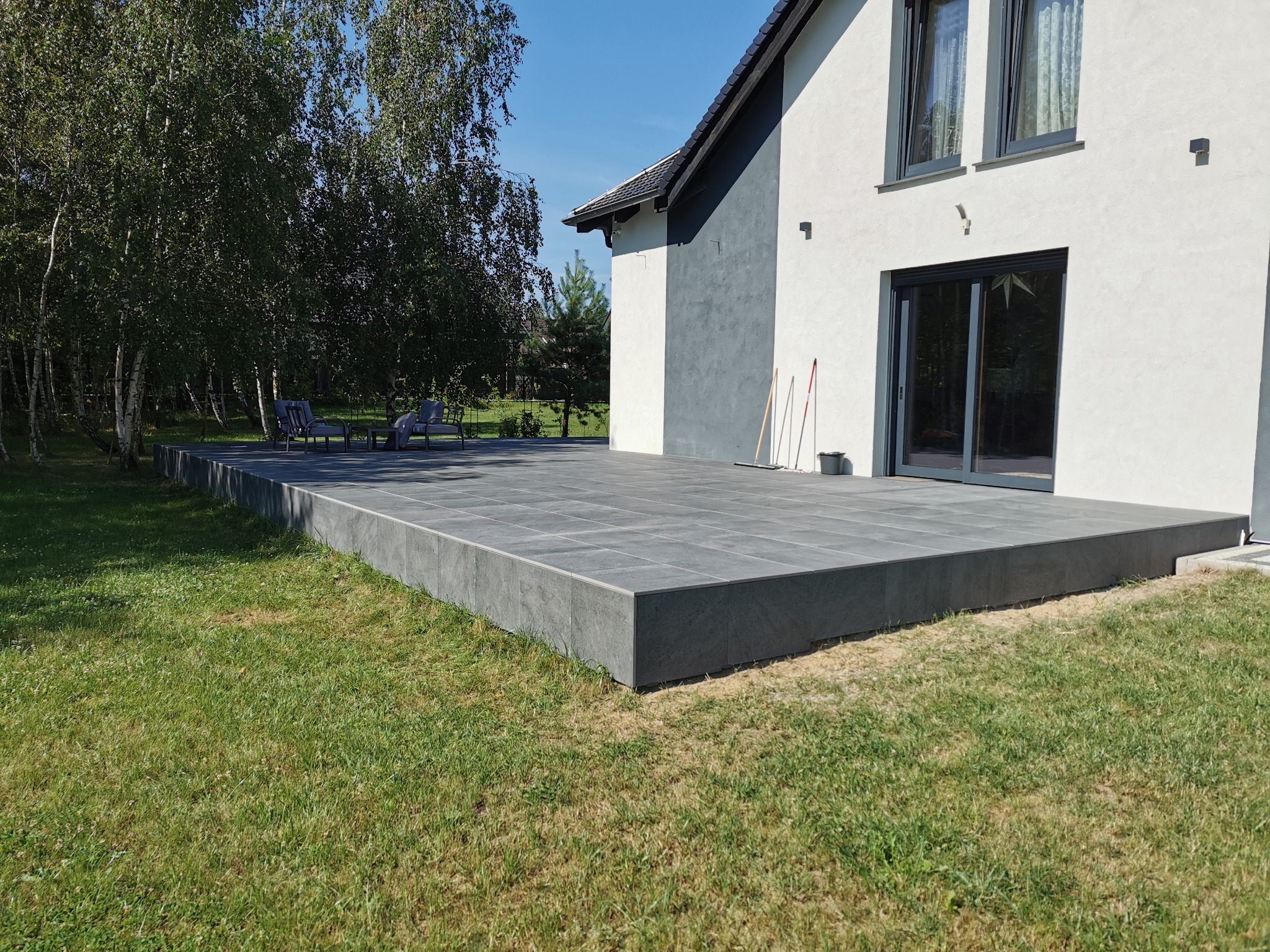 Nowoczesny taras z szarych płyt kamiennych przylegający do jasnego domu z ciemnymi ramami okiennymi, widoczne drzewa w tle i zestaw mebli ogrodowych.