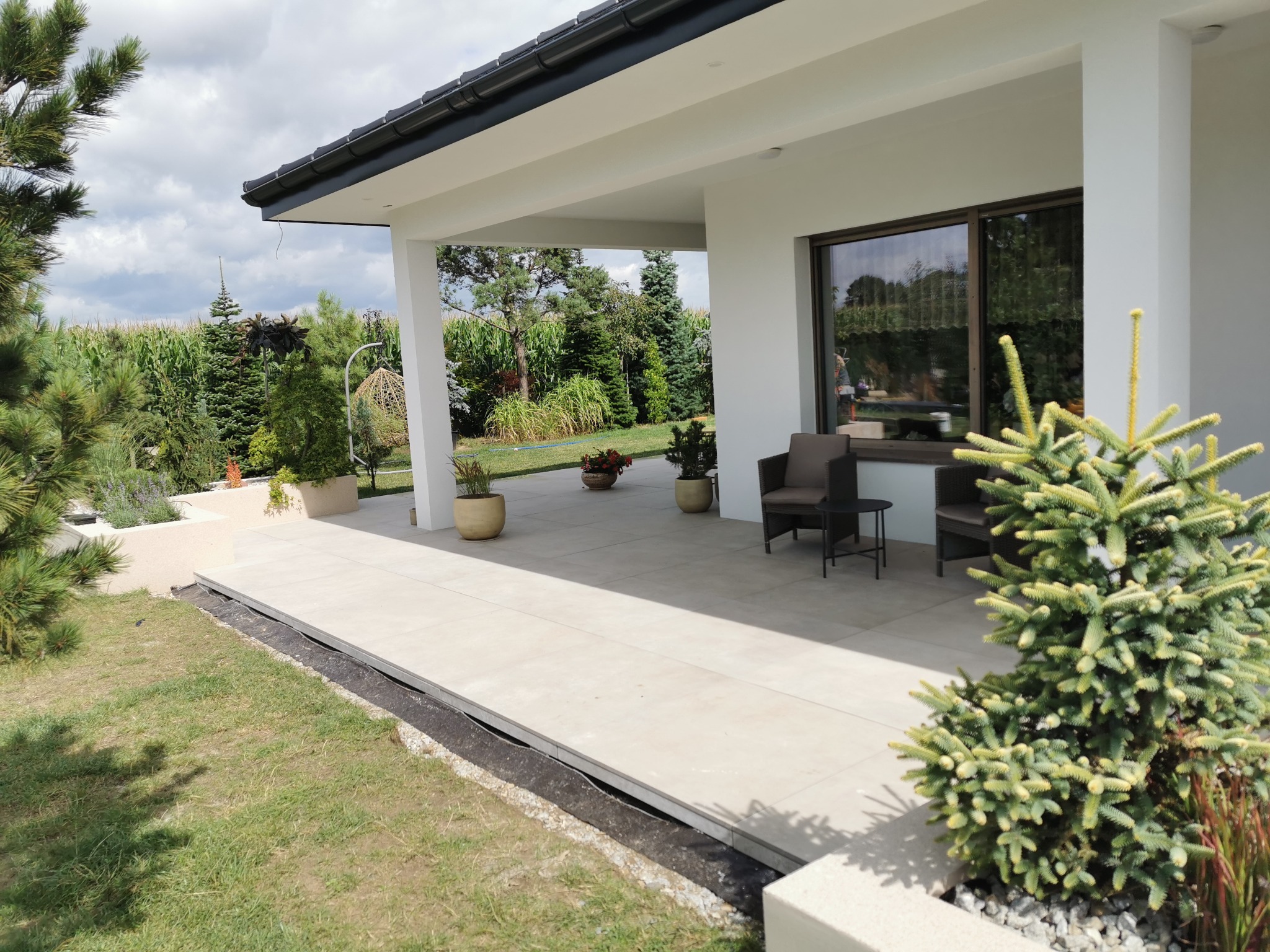 Nowoczesny taras z jasnych płyt z odpływem liniowym, widoczne meble ogrodowe i dekoracyjne rośliny.