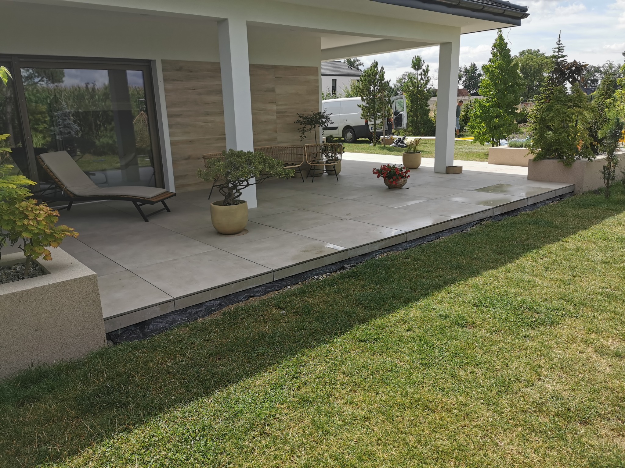 Nowoczesny taras z jasnych, kwadratowych płyt, przylegający do domu z dużymi oknami i zadaszeniem, widoczne meble ogrodowe, rośliny w donicach i trawnik.