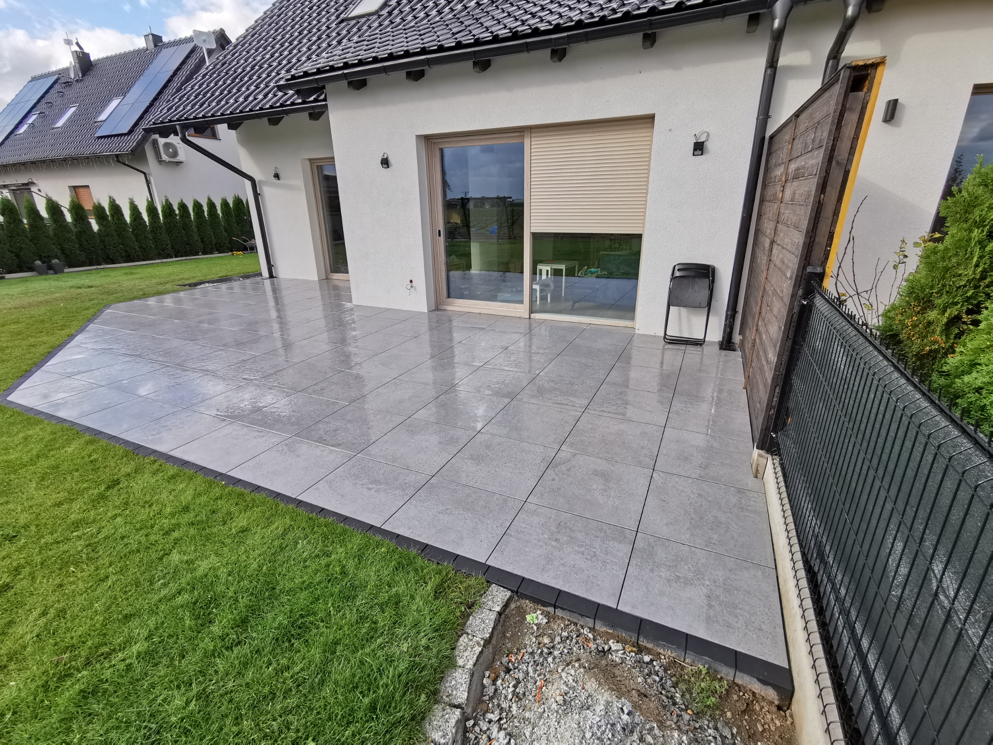 Nowoczesny, szary taras z płytek, obrzeża z ciemnego kamienia, przylegający do domu z dużymi przesuwnymi drzwiami i roletą, obok trawnik z zieloną trawą.