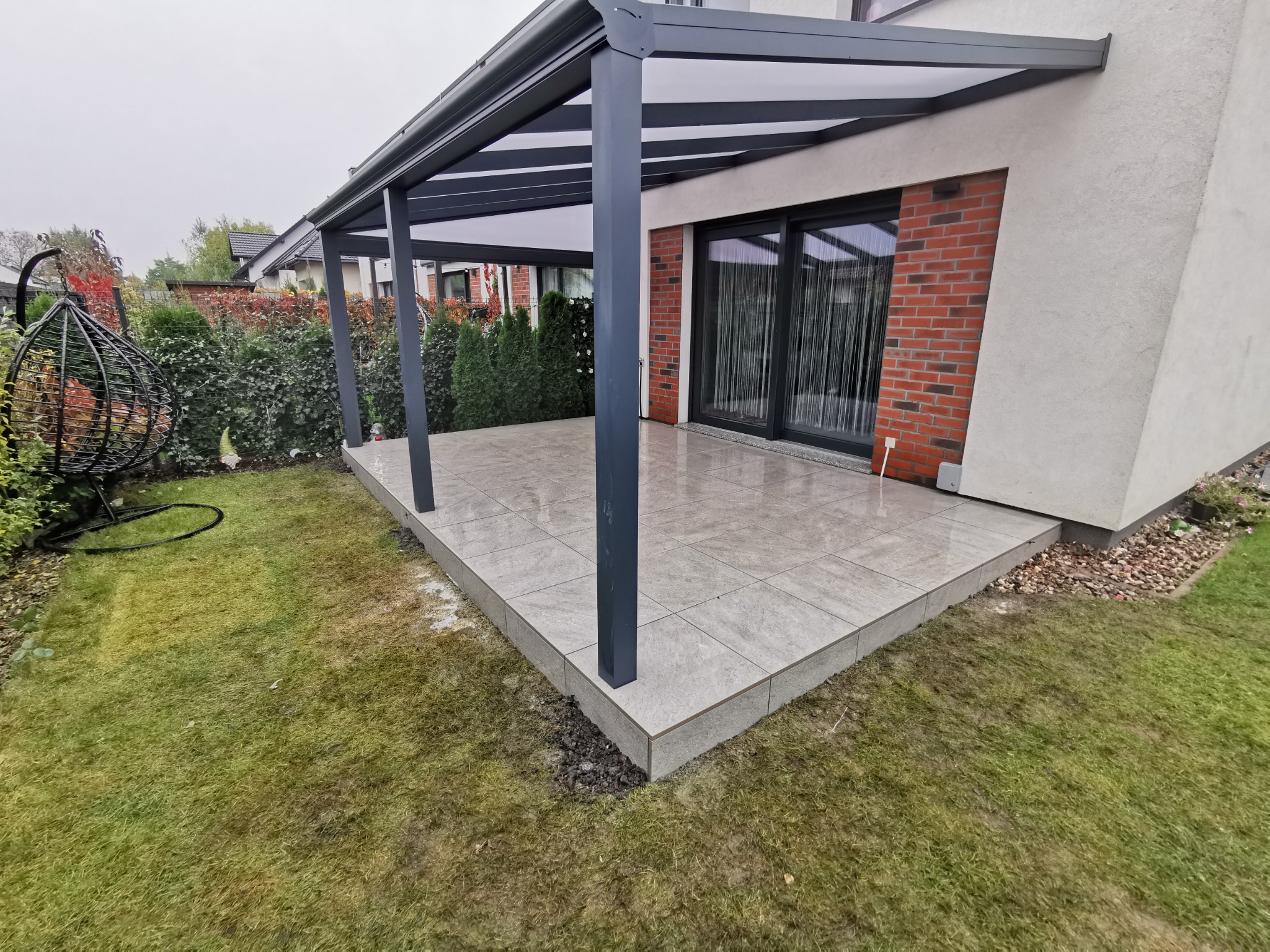Zadaszony taras z szarych płyt kamiennych, widok na dom z ceglaną elewacją i czarnymi oknami przesuwnymi, obok trawnik i wiszący fotel.