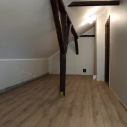 Pomalowane poddasze z drewnianą konstrukcją, nową podłogą i widocznymi przewodami elektrycznymi przy ścianie.