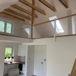 Jasne wnętrze domu z antresolą, widoczne drewniane belki stropowe, szklane balustrady antresoli, biała kuchnia z czarnymi akcentami i schody z drewnianą poręczą.