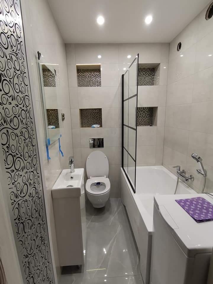 Nowoczesna łazienka z szarymi płytkami, zabudowaną wanną z czarną szybą, toaletą i umywalką na szafce. Na ścianie dekoracyjne wnęki z mozaiką i oświetleniem LED.