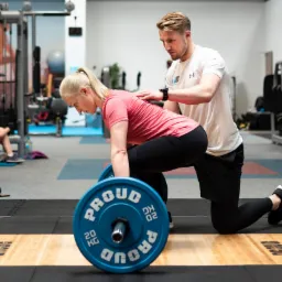 Kobieta w różowej koszulce wykonuje martwy ciąg z niebieskim obciążeniem pod okiem trenera w białej koszulce, który koryguje jej postawę w jasnej siłowni.