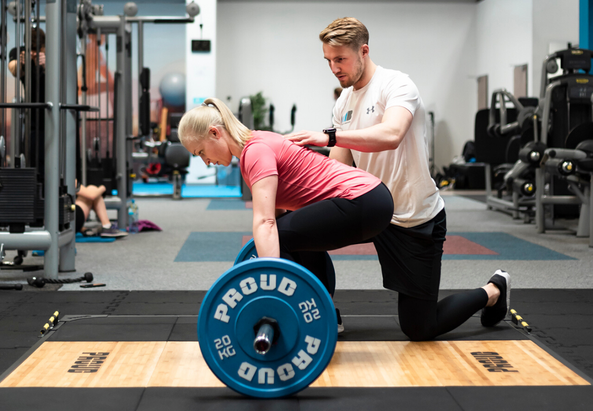 Kobieta w różowej koszulce wykonuje martwy ciąg z niebieskim obciążeniem pod okiem trenera w białej koszulce, który koryguje jej postawę w jasnej siłowni.