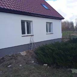 Świeżo otynkowany, biały dom jednorodzinny w Bartoszycach z czerwoną dachówką i szarym cokołem, widoczna drabina oparta o ścianę, sugerująca trwające prace wykończeniowe.