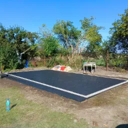 Wykończona, prostokątna powierzchnia z ciemnej kostki brukowej z jasną obwódką, widok z góry na tle zieleni ogrodowej i błękitnego nieba, butelka wody mineralnej na trawie w lewym dolnym rogu.