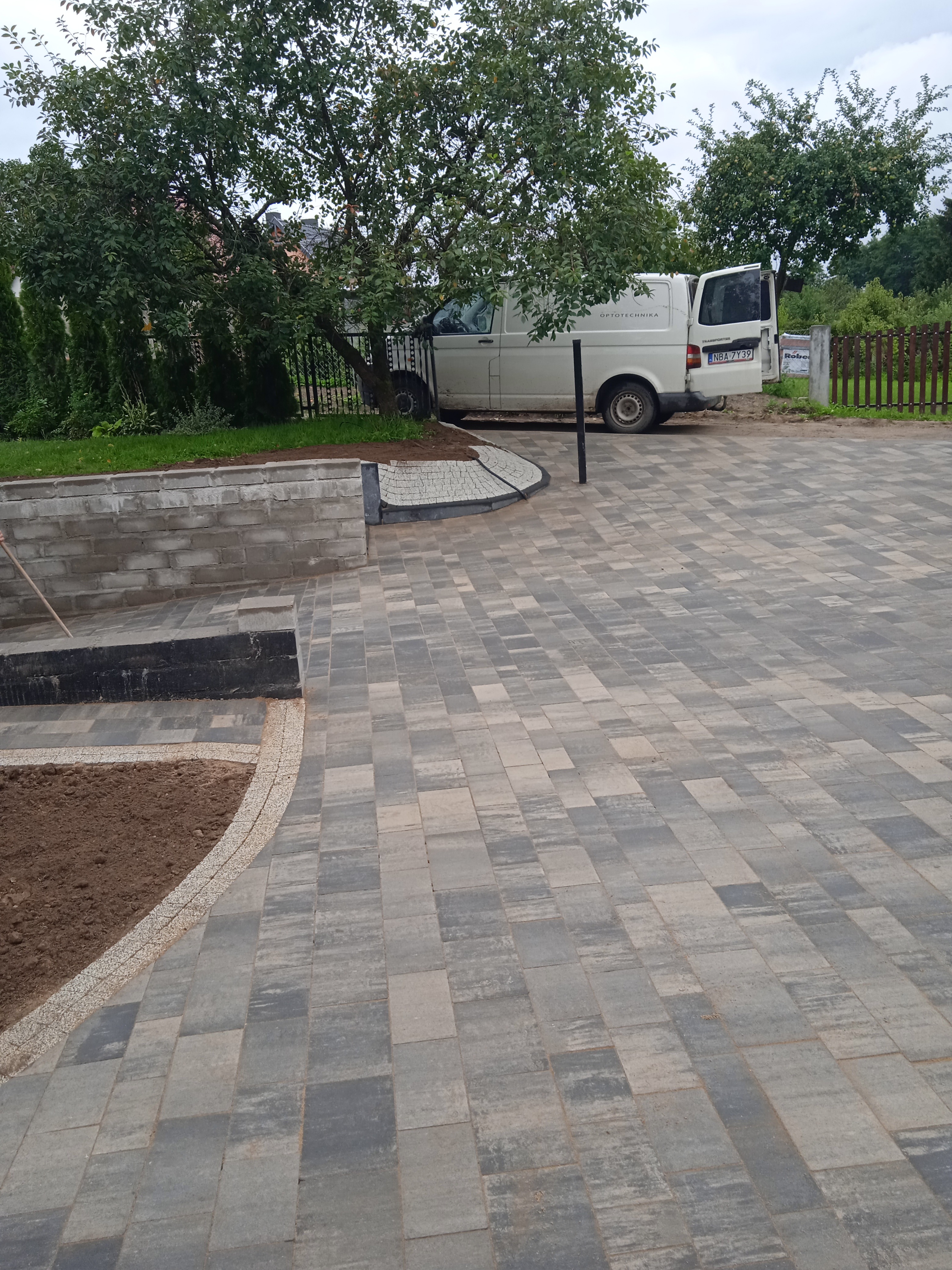 Ułożona kostka brukowa w odcieniach szarości z obrzeżem z drobnych kamieni, widoczny fragment rabaty z ziemią, w tle zaparkowany biały bus z otwartymi drzwiami.