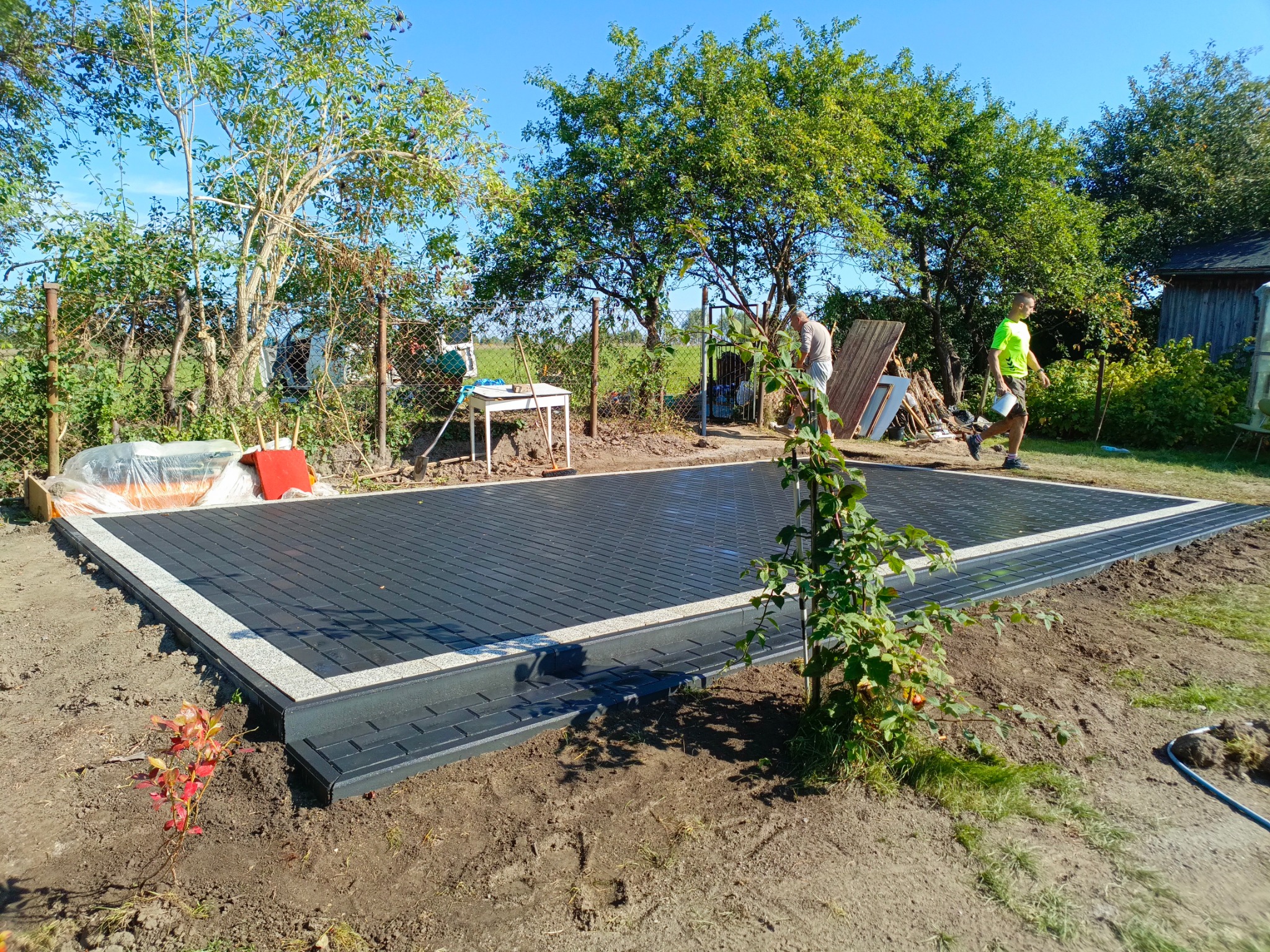 Nowoczesna, ciemnoszara nawierzchnia z kostki brukowej z jasnoszarym obramowaniem, częściowo ułożona na przygotowanym terenie, z widocznymi pracownikami w tle i elementami zagospodarowania terenu.