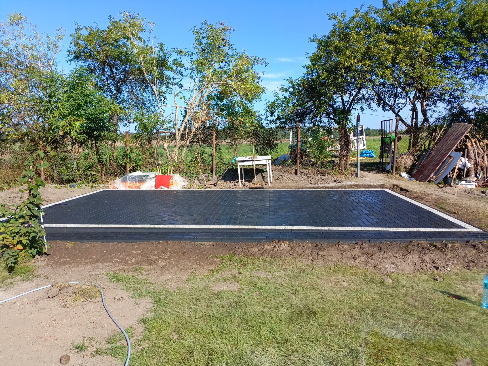 Nowo ułożona, prostokątna nawierzchnia z ciemnej kostki brukowej z jasnym obrzeżem, otoczona ziemią i trawą, na tle drzew i błękitnego nieba.