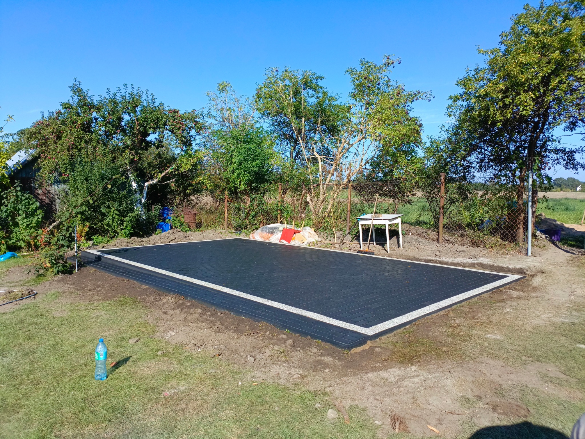 Wykończona, prostokątna powierzchnia z ciemnej kostki brukowej z jasną obwódką, widok z góry na tle zieleni ogrodowej i błękitnego nieba, butelka wody mineralnej na trawie w lewym dolnym rogu.