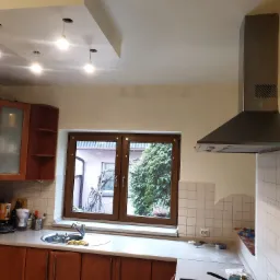 Kuchnia z drewnianymi szafkami, widok na okno z zielenią, szary okap, białe płytki na ścianie i nowoczesne oświetlenie sufitowe.