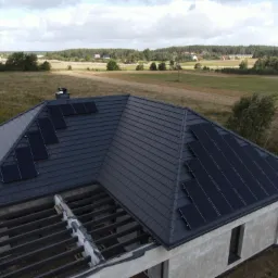 Montaż paneli fotowoltaicznych na dachu domu w trakcie budowy, widoczne krokwie i dachówka w kolorze grafitowym, w tle pole i zabudowania.