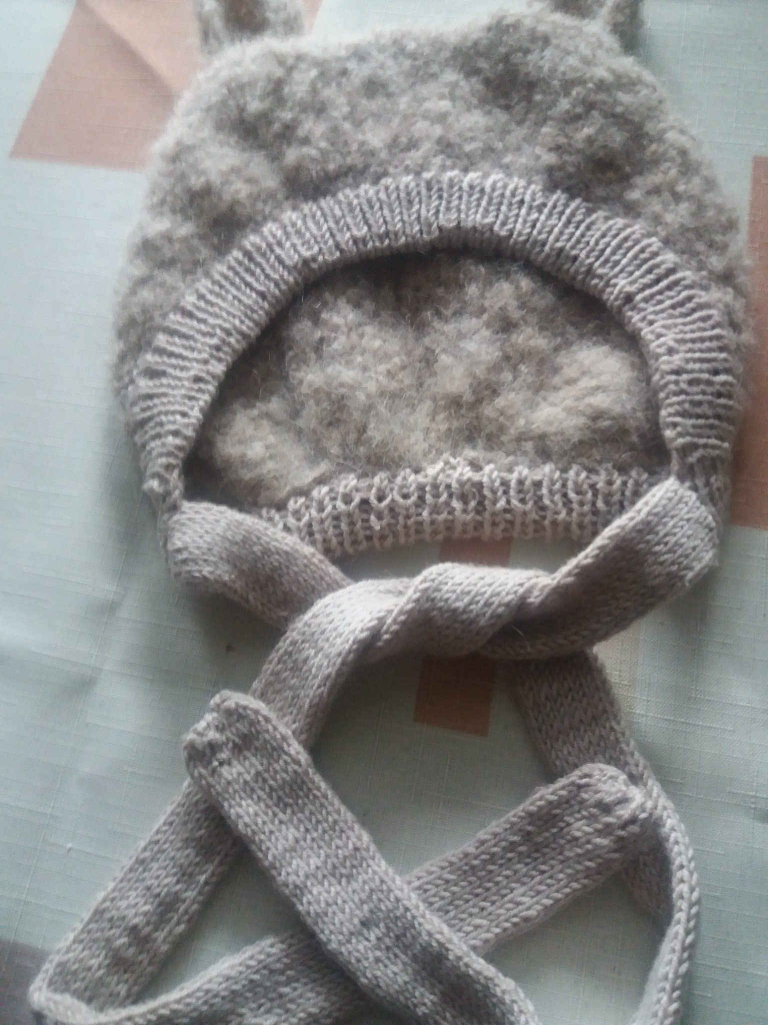 Ręcznie robiona, szara, wełniana czapka z uszami, leżąca na wzorzystym tle.