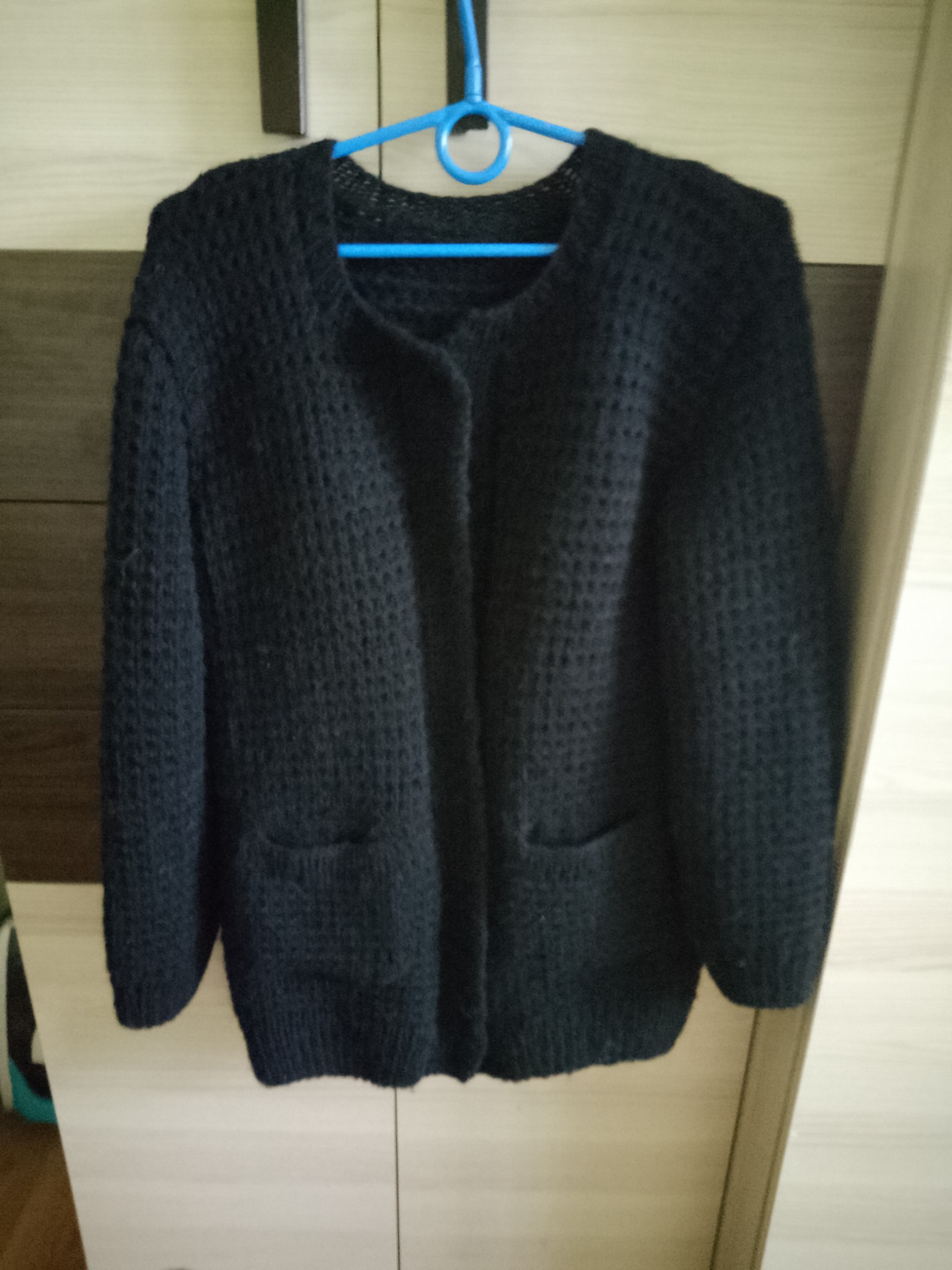 Czarny, ręcznie robiony kardigan z grubym splotem, wiszący na niebieskim wieszaku na tle szafy z panelami w odcieniach beżu i brązu.