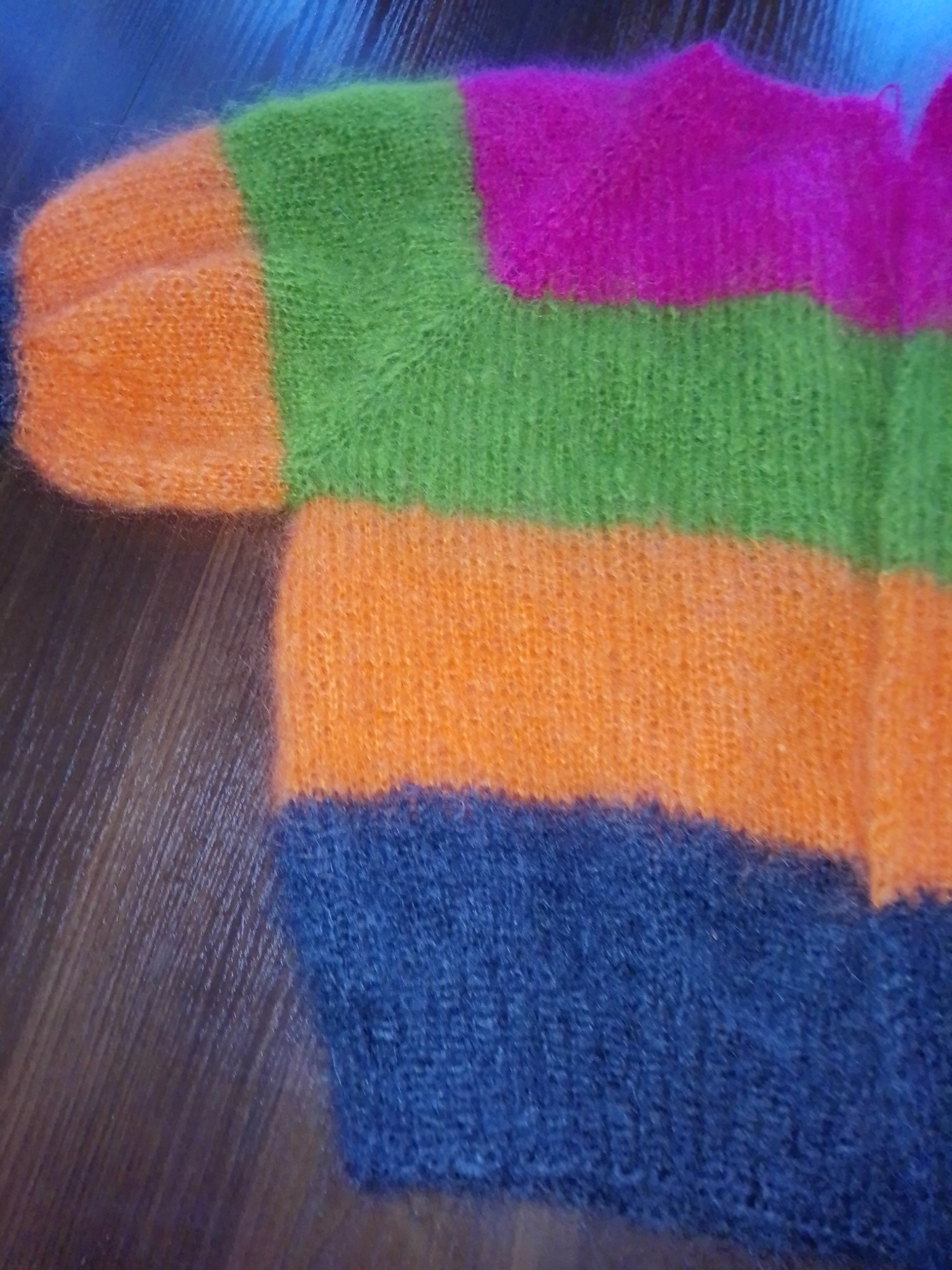 Fragment puszystego, ręcznie robionego swetra w poziome pasy: pomarańczowy, zielony, fuksja, pomarańczowy i granatowy, leżącego na drewnianej powierzchni.