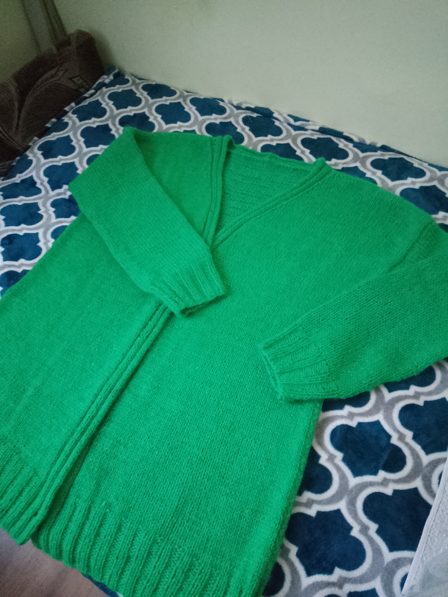 Jasnozielony, ręcznie robiony kardigan z dekoltem w serek leży na wzorzystej narzucie, prezentując detale splotu i wykończenia.