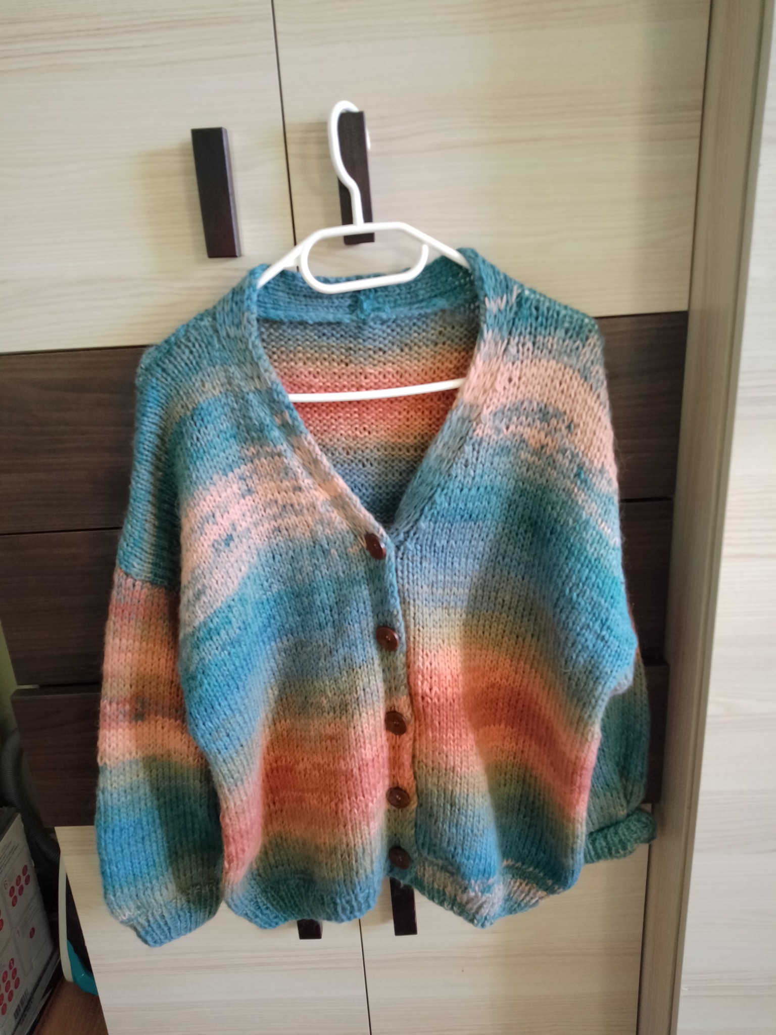 Ręcznie robiony, rozpinany sweter z grubych włóczek w odcieniach błękitu, pomarańczy i różu, wiszący na białym wieszaku na tle szafy z drewnianymi elementami.