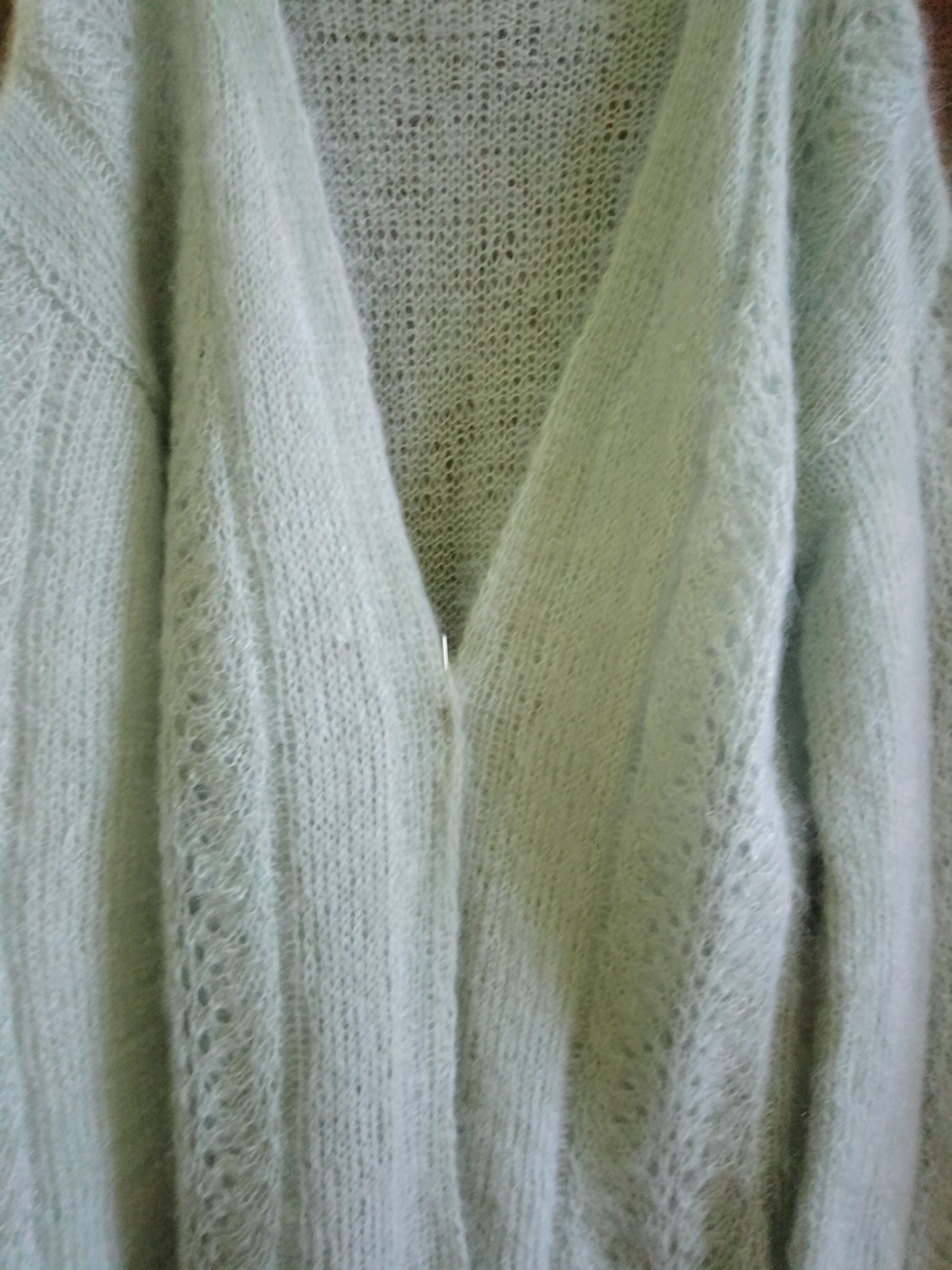 Delikatny, ażurowy kardigan damski w odcieniu pastelowej zieleni, zapinany na jeden srebrny guziczek, wykonany z miękkiej włóczki moherowej o widocznej strukturze splotu.