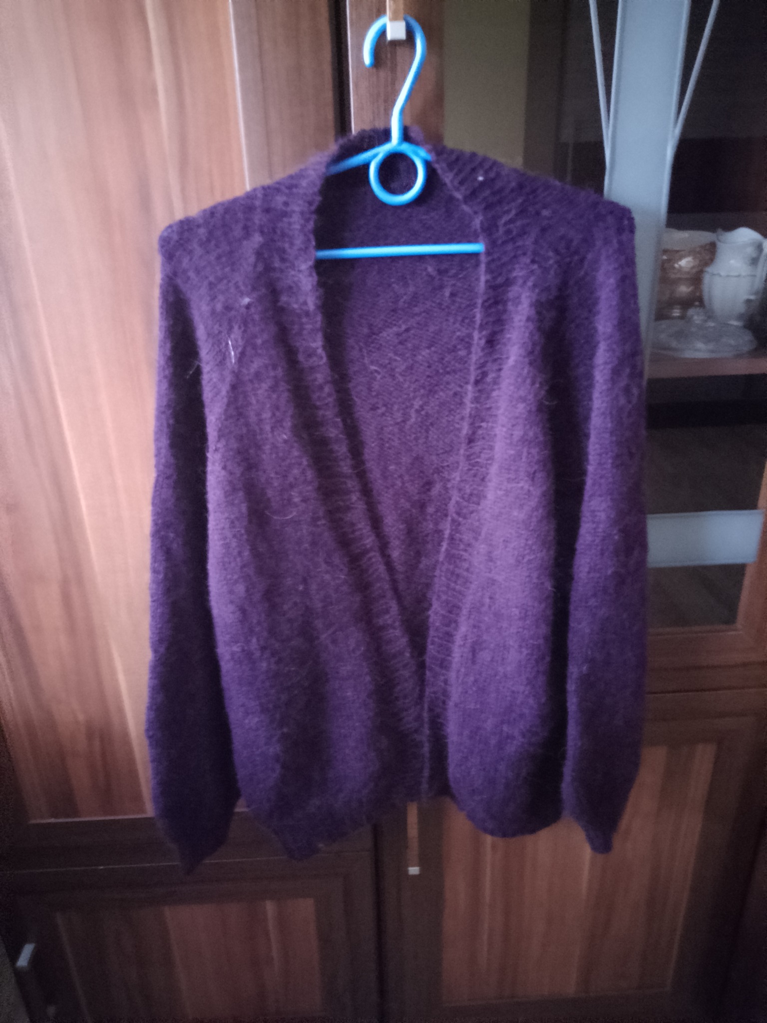Fioletowy, włochaty kardigan z dzianiny, wiszący na niebieskim wieszaku na tle drewnianych drzwi szafy w Jednorożcu.