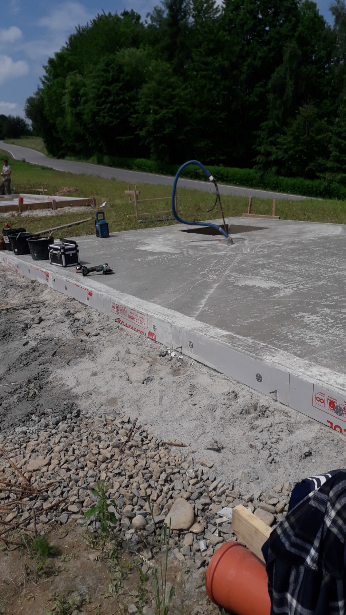 Wylany fundament z izolacją obwodową, widoczne narzędzia budowlane, niebieski wąż do pompowania betonu, teren budowy z gruzem i fragmentem drogi w tle.