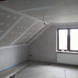 Poddasze w trakcie montażu płyt gipsowo-kartonowych, widoczne łączenia płyt zaszpachlowane, surowa żarówka zwisa z sufitu, okno z grzejnikiem w tle.