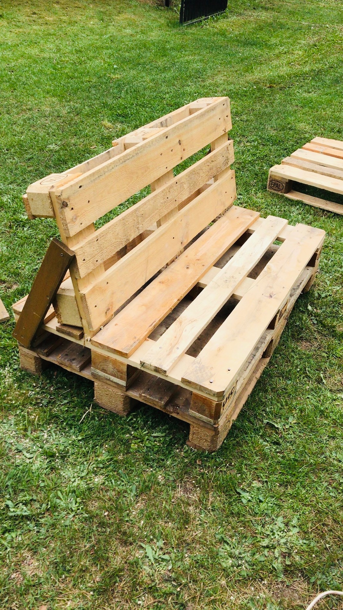 Kreatywny fotel ogrodowy wykonany z europalet, ustawiony na trawniku. Widoczna konstrukcja oparcia i siedziska. W tle kolejna paleta.